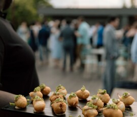 Server with a tray of herb-garnished appetizers at a Gulf South outdoor wedding, crowd blurred in background.