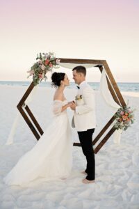 Bride and groom barefoot on sandy beach, holding hands by a floral hexagonal arch with ocean and pastel sky behind.