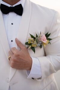 Person in white tuxedo with black bow tie and gold ring adjusts lapel with pink and yellow boutonniere at New Orleans wedding.