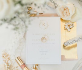 Elegant wedding flat lay: gold-detailed invitation, rings, white roses, shoes, lipstick, pearl hair accessories, earrings.