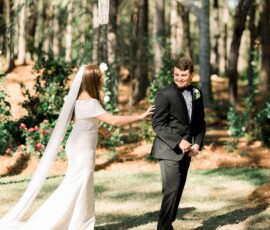 Bride in white dress and veil touches smiling groom’s shoulder outdoors amid greenery at their Gulf South wedding.