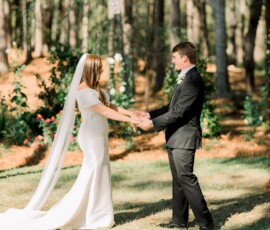 Bride and groom hold hands in a sunlit Louisiana forest clearing during their wedding, bride in white dress, groom in black suit.