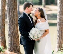 Bride and groom smile at each other by trees and a pond during their Louisiana wedding; bride in white, groom in black suit.
