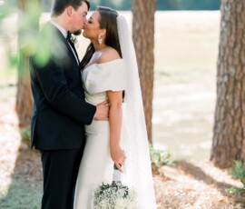 Bride and groom kiss among trees at their Louisiana wedding, sunlight filtering as she holds white flowers in her white gown.