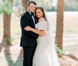 Bride in white dress and groom in black tuxedo embrace outdoors, smiling among tall trees with sunlight filtering through.