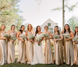 Bride in white and eight bridesmaids in gold pose with bouquets by a white barn on a sunny Gulf South wedding day.
