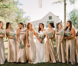 Bride in white poses with eight bridesmaids in gold dresses, all smiling with bouquets by a white barn on a sunny day.