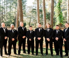 Eight men in black tuxedos stand outdoors among tall trees with sunlight filtering through, smiling at the camera.