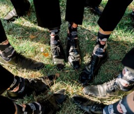 People in black dress shoes and pants stand in a circle, showing off socks with a face and “Here Comes the Groom” text.