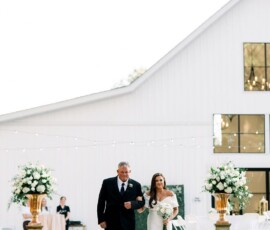 Bride in white dress walks with older man by a white barn and flower arrangements, inspiring elegant wedding ideas.