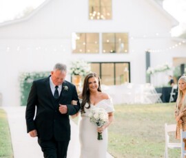 Bride in off-the-shoulder gown walks with older man outside white barn at elegant New Orleans wedding.