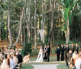 Bride and groom at an outdoor forest altar, surrounded by wedding party and guests, sunlight streaming through the trees.