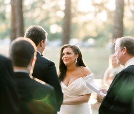 Bride and groom exchange vows outdoors among trees and sunlight, surrounded by guests during a heartfelt wedding ceremony.