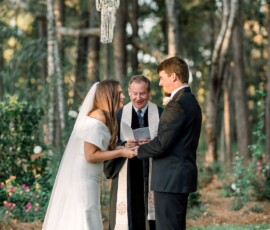 Bride and groom smiling and holding hands at an outdoor New Orleans wedding, officiant behind them, trees in background.