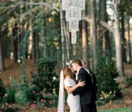 Bride and groom kiss beneath a crystal chandelier outdoors, with trees and greenery at a Gulf South wedding.