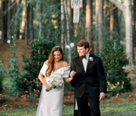 Bride and groom walk hand in hand outdoors, smiling beneath a chandelier, surrounded by trees and flowers at a Gulf South wedding.