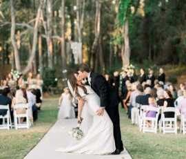 Bride and groom kiss on an outdoor aisle in the woods, surrounded by guests and their wedding party in the background.