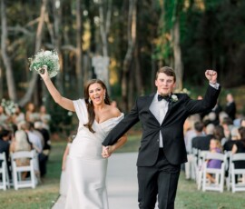 Bride and groom walk hand in hand outdoors after a New Orleans wedding, celebrating as guests watch among trees.