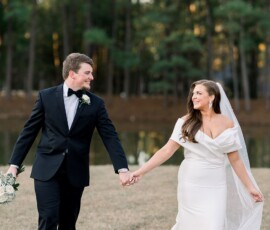 Bride in white dress and groom in black tuxedo walk hand-in-hand on grass by trees and a pond at a Louisiana wedding.