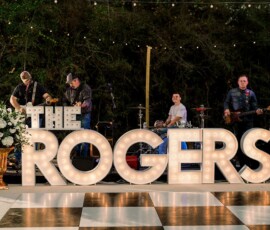 A band plays on stage behind lit “THE ROGERS” letters, floral décor, and string lights on a checkered wedding dance floor.