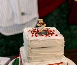 Square two-tier white iced cake with red velvet layers, bear topper, red crumbs, and blurred baseball jersey in the background.