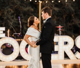 Bride and groom dance outdoors at night amid glowing letters and string lights, capturing a joyful wedding moment.