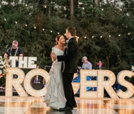 Bride and groom dance on a checkered floor at their Louisiana wedding, THE ROGERS lit up behind them amidst flowers and musicians.