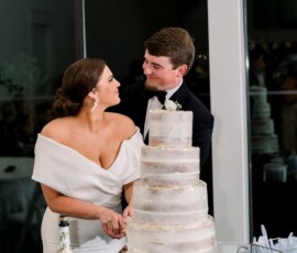 Bride and groom cut a three-tiered cake at an elegant wedding, smiling beside white flowers and champagne.