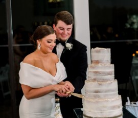 Bride and groom cut a tall white wedding cake, smiling together at an elegant Louisiana wedding with white flowers on the table.