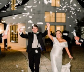 Smiling newlyweds walk at night through falling confetti, hand-in-hand outside a brightly lit Louisiana wedding venue.