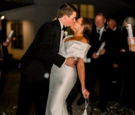 Bride and groom share a passionate kiss at night, surrounded by guests and confetti, with a glowing house in the background.