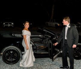 Bride in white dress smiles by open car door as groom in tuxedo opens it, capturing Gulf South wedding elegance at night.