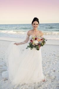 Bride in off-shoulder white dress, barefoot on a sandy beach with bouquet, ocean, and pastel sky—Louisiana wedding inspiration.