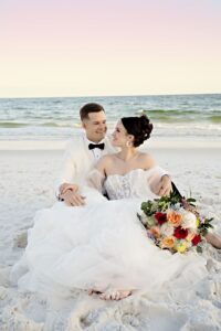 Bride and groom sit on a sandy beach at sunset, with bouquet, white attire, ocean views, and pastel Gulf South sky.