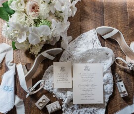 Gulf South bridal accessories on wood floor: white heels, bouquet, lace, perfume, invites, jewelry, cufflinks, monogram cloth.