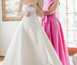 A bride in a white gown smiles as a woman in pink helps her get ready in a bright Gulf South wedding room.