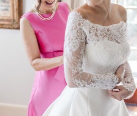 A woman in a pink dress helps a smiling bride in a white lace gown prepare by a window before their New Orleans wedding.