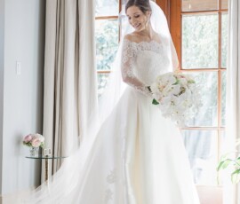 Bride in white gown and veil stands by sunny window with cascading bouquet, smiling before Louisiana wedding ceremony.