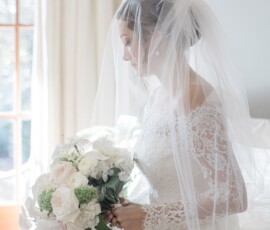 Bride in lace dress and veil holds white and blush bouquet, softly lit by window, facing right at classic Louisiana wedding.