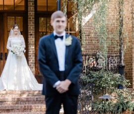 Bride in white smiles on brick steps behind out-of-focus groom; New Orleans wedding with lush greenery and large windows.