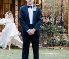 Bride in white gown and veil approaches groom in tuxedo for a first look outside a brick house at Louisiana wedding.