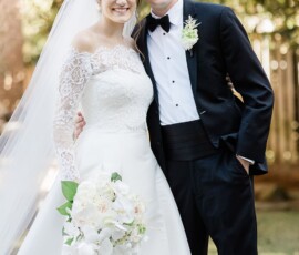Bride in lace gown and groom in black tuxedo smile outdoors with white bouquet, framed by trees—Gulf South wedding inspiration.