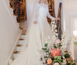 Bride in long-sleeve white gown and veil on staircase, smiling with bouquet of pink and white flowers—Gulf South wedding inspiration.