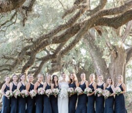 Bride in white with twelve bridesmaids in dark dresses, holding bouquets under sunlit trees—New Orleans wedding inspiration.