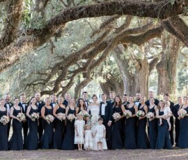 Wedding party stands smiling under arching trees in Louisiana, with bridesmaids in black dresses and groomsmen in black suits.