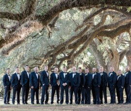 Sixteen men in tuxedos pose outdoors under sprawling tree branches for a formal group wedding photo, inspired by New Orleans elegance.