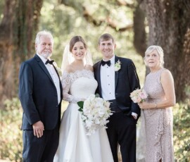 Bride and groom with an older couple smile outdoors at a Louisiana wedding, surrounded by trees and greenery.