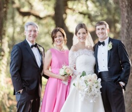 Four people smile outdoors at a wedding: bride, groom, woman in pink with flowers, and older man in tuxedos.
