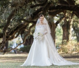 Bride in a white dress and veil stands under large trees, holding a white bouquet and smiling—a Louisiana wedding moment.
