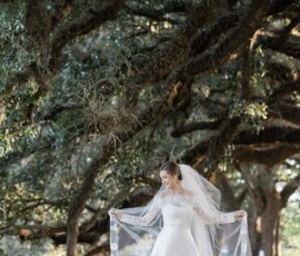 Bride in white dress and veil stands under sprawling Louisiana tree, softly smiling as sunlight filters through leaves.